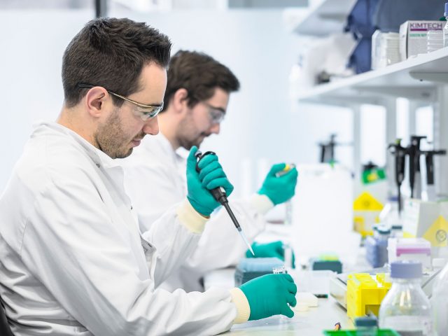 People wearing lab coats, using pipettes to drop liquid into test tubes.