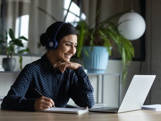 Woman with headset on taking notes during a video call on her laptop.