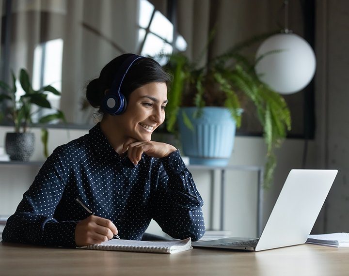 Woman with headset on taking notes during a video call on her laptop.