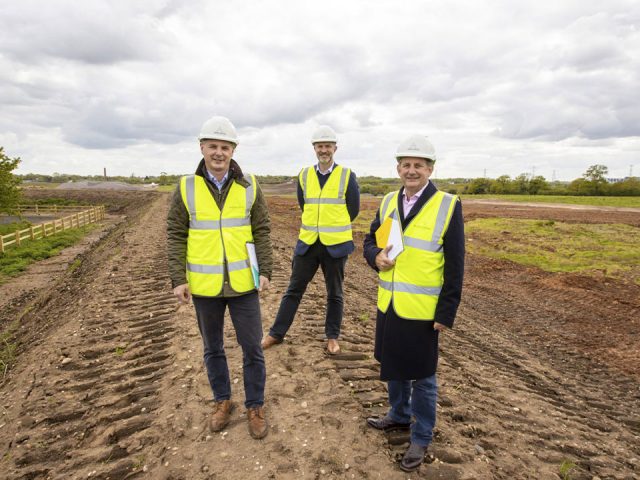 Men in hi-vis jackets and hard hats standing in a field. Digger tracks are visible under their feet.