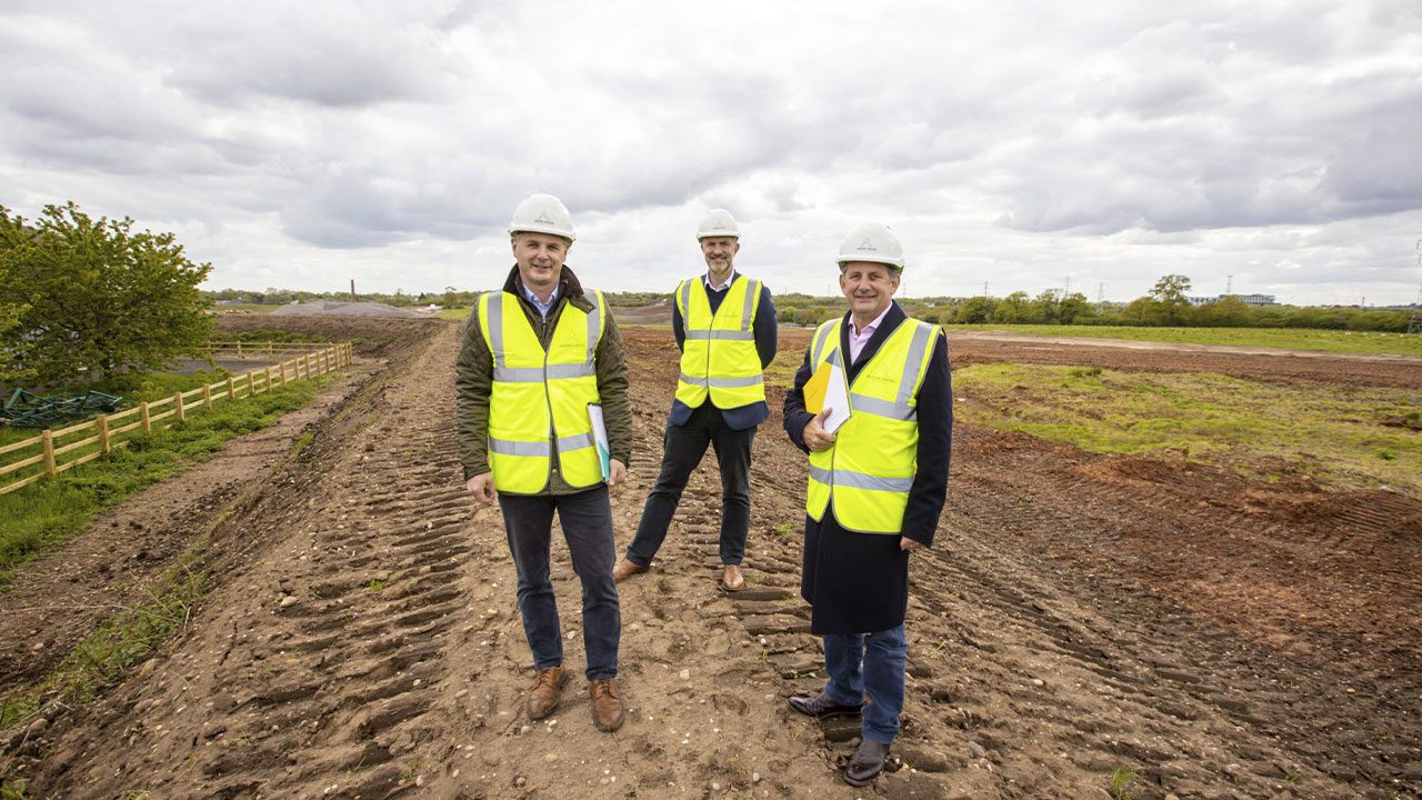 Men in hi-vis jackets and hard hats standing in a field. Digger tracks are visible under their feet.
