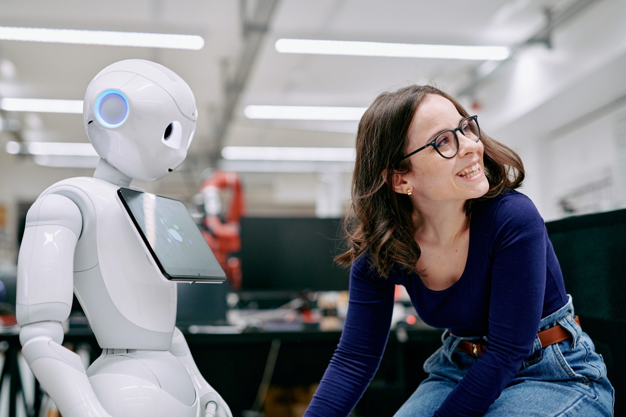 A woman smiling next to a white robot with a tablet computer attached to it's chest.