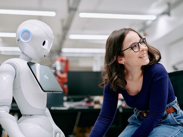 A woman smiling next to a white robot with a tablet computer attached to it's chest.