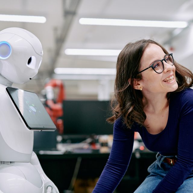 A woman smiling next to a white robot with a tablet computer attached to it's chest.