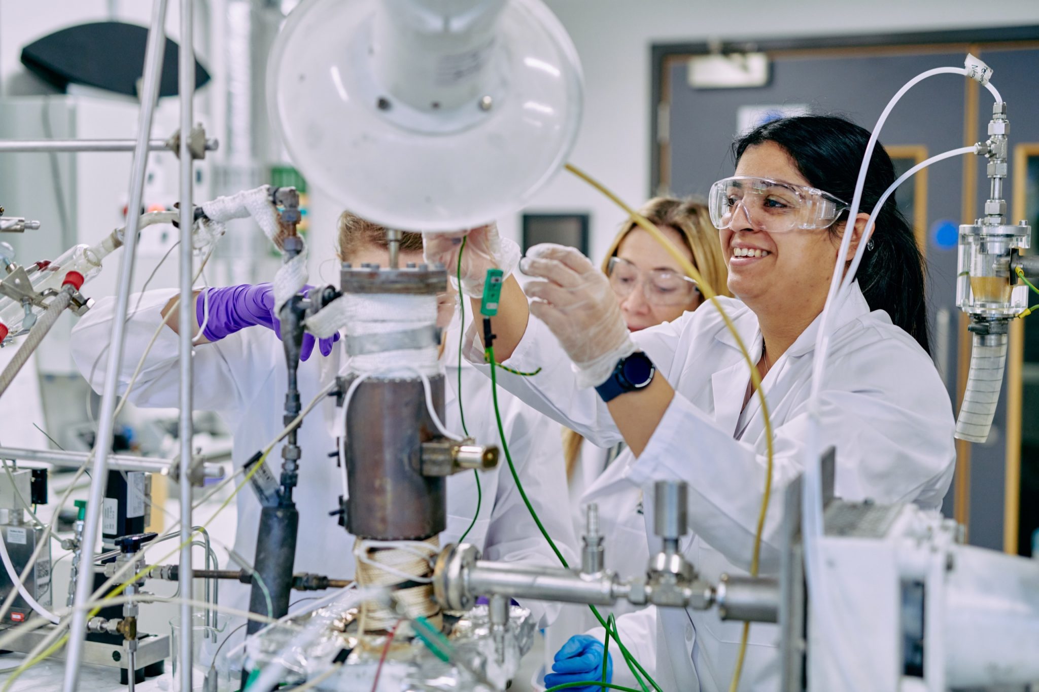 People wearing lab coats and safety goggles connecting machinery on a desk.