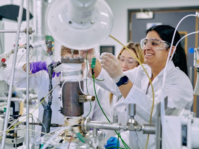 People wearing lab coats and safety goggles connecting machinery on a desk.