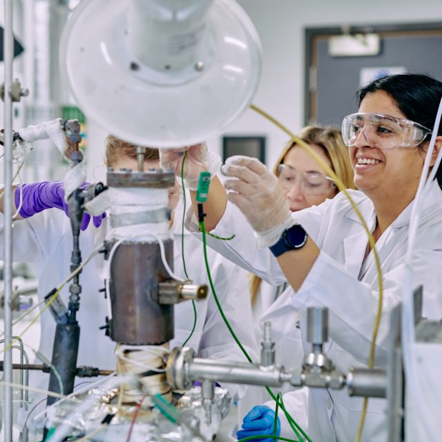 People wearing lab coats and safety goggles connecting machinery on a desk.