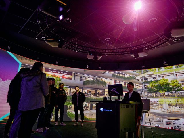 People gathered around a speaker giving a talk using a screen. They are in a multi-level shopping centre with hanging plants trailing over the balconies.