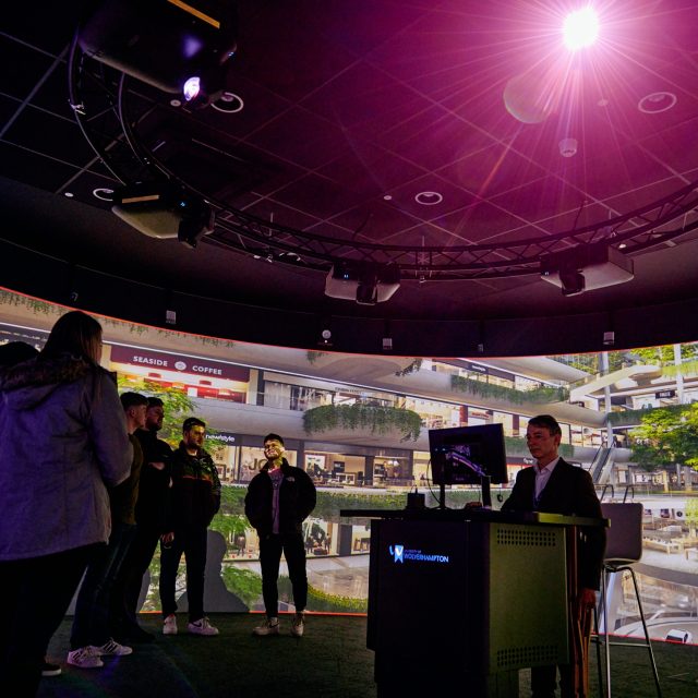 People gathered around a speaker giving a talk using a screen. They are in a multi-level shopping centre with hanging plants trailing over the balconies.