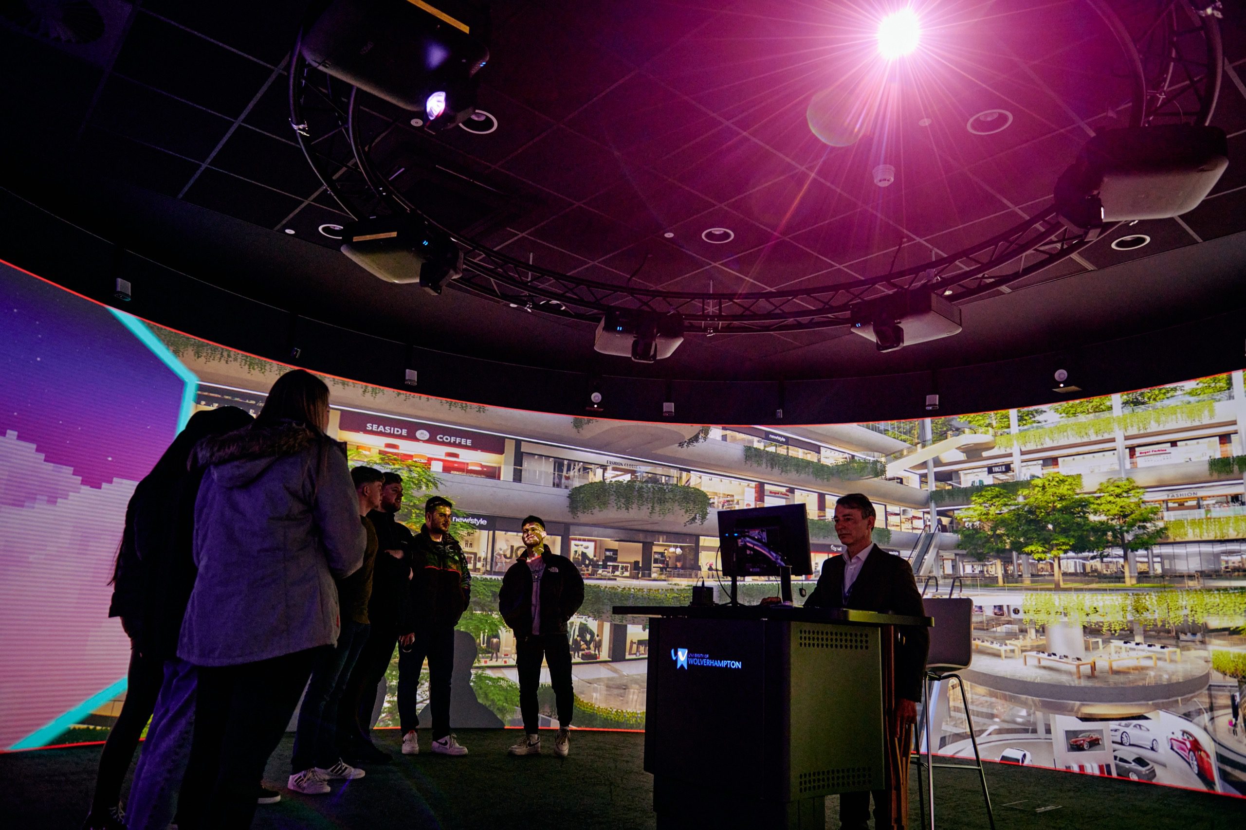 People gathered around a speaker giving a talk using a screen. They are in a multi-level shopping centre with hanging plants trailing over the balconies.