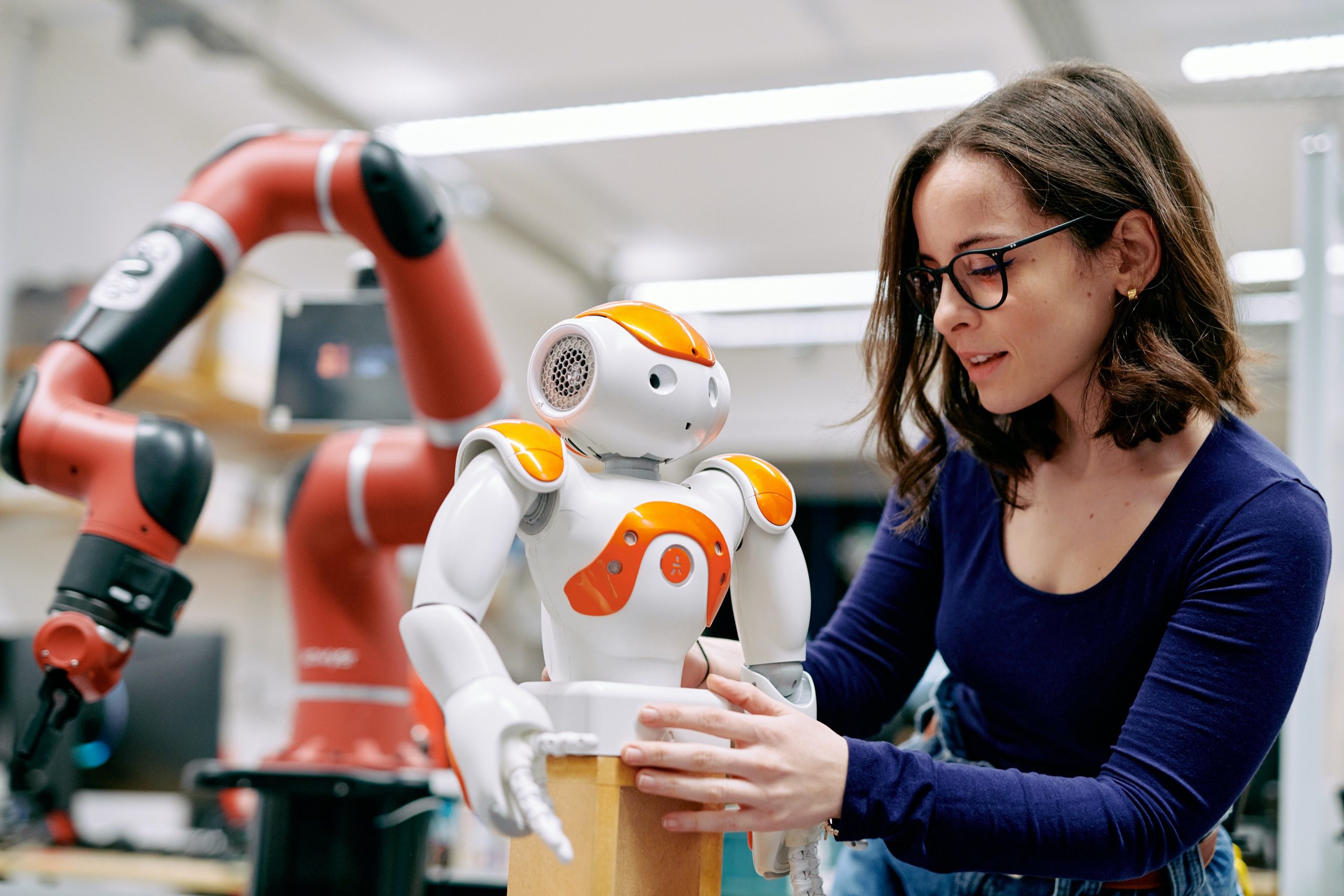 A woman placing a small robot onto a platform. The robot only has an upper body and arms and is white with orange accents.