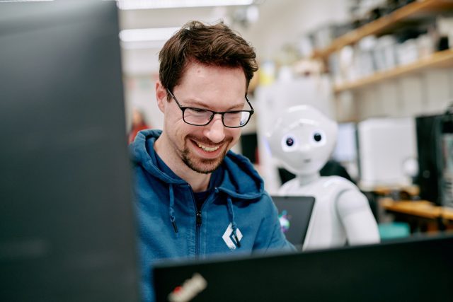 A man smiling at a laptop. Behind him a white robot with glowing blue eyes is visible.