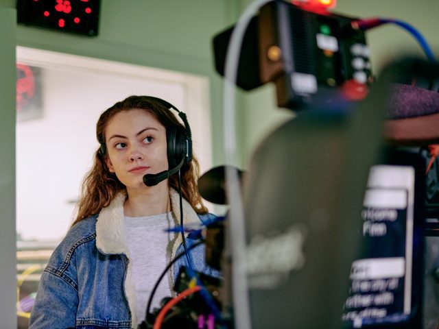 A woman wearing a headset with microphone. Blurry cameras are visible in the foreground.