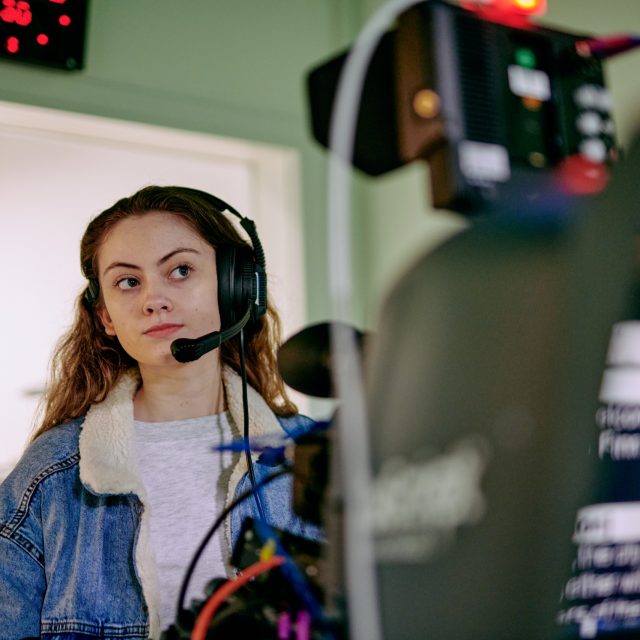 A woman wearing a headset with microphone. Blurry cameras are visible in the foreground.