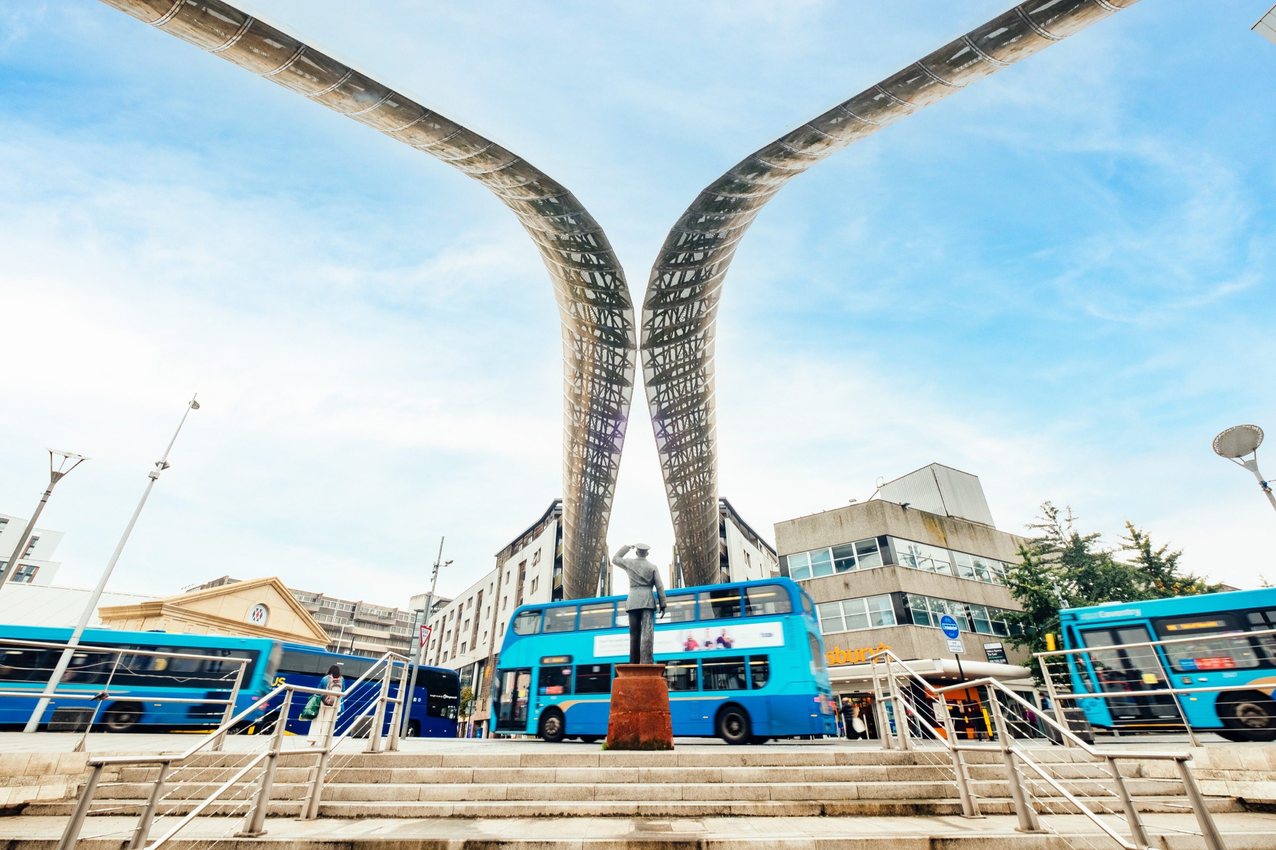 Statue of a person saluting in military wear on a busy road. Overhead 2 metal arches curve towards each other but don't meet.