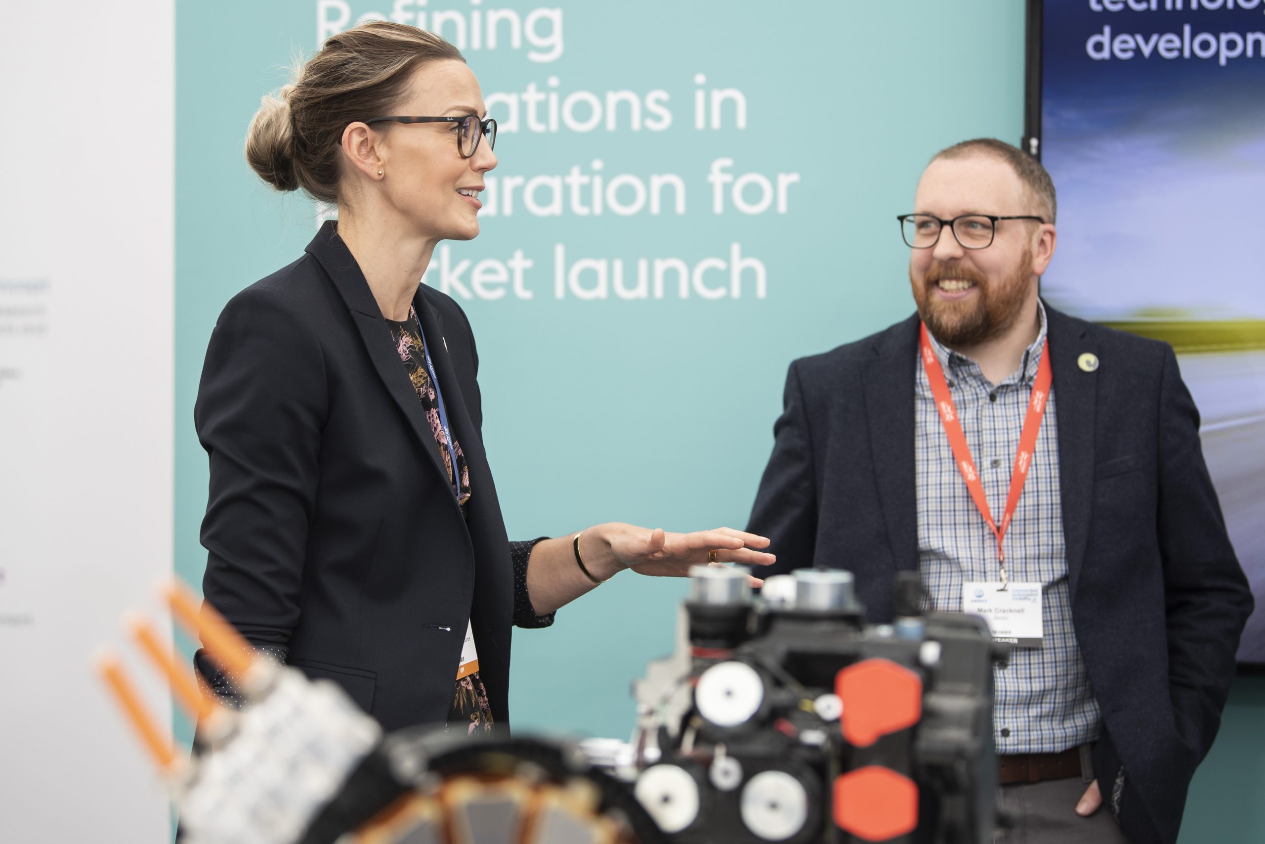 People talking with conference lanyards on. Part of an engine is visible but blurred in front of them.