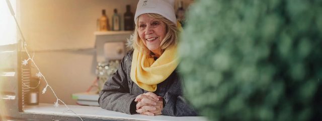 Woman wearing a scarf and beanie hat with her hands clasped, leaning on a counter.