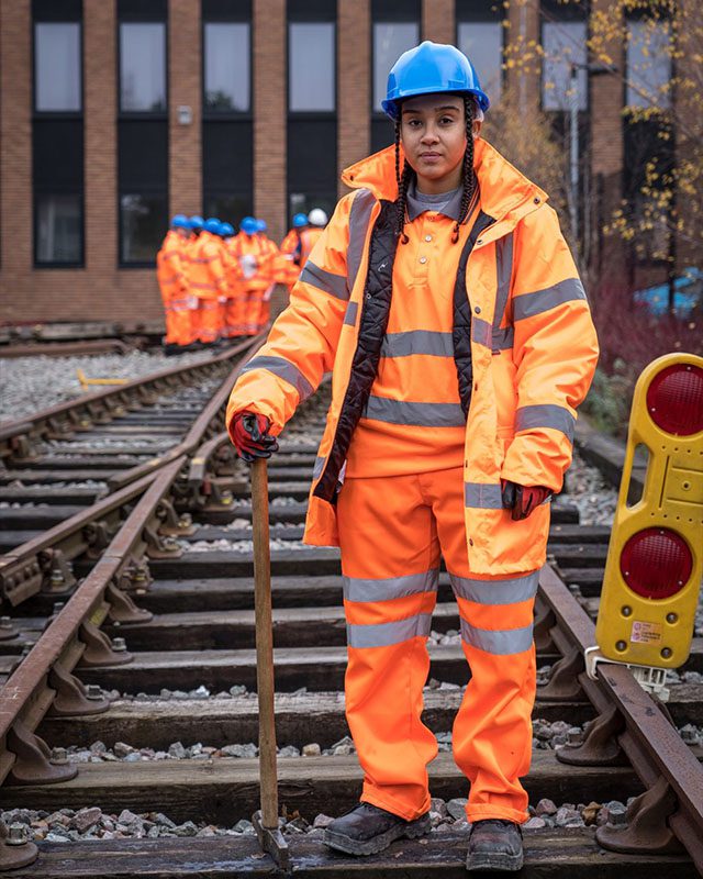 A rail worker standing on railway tracks, wearing an orange hi-vis uniform and a hard hat.