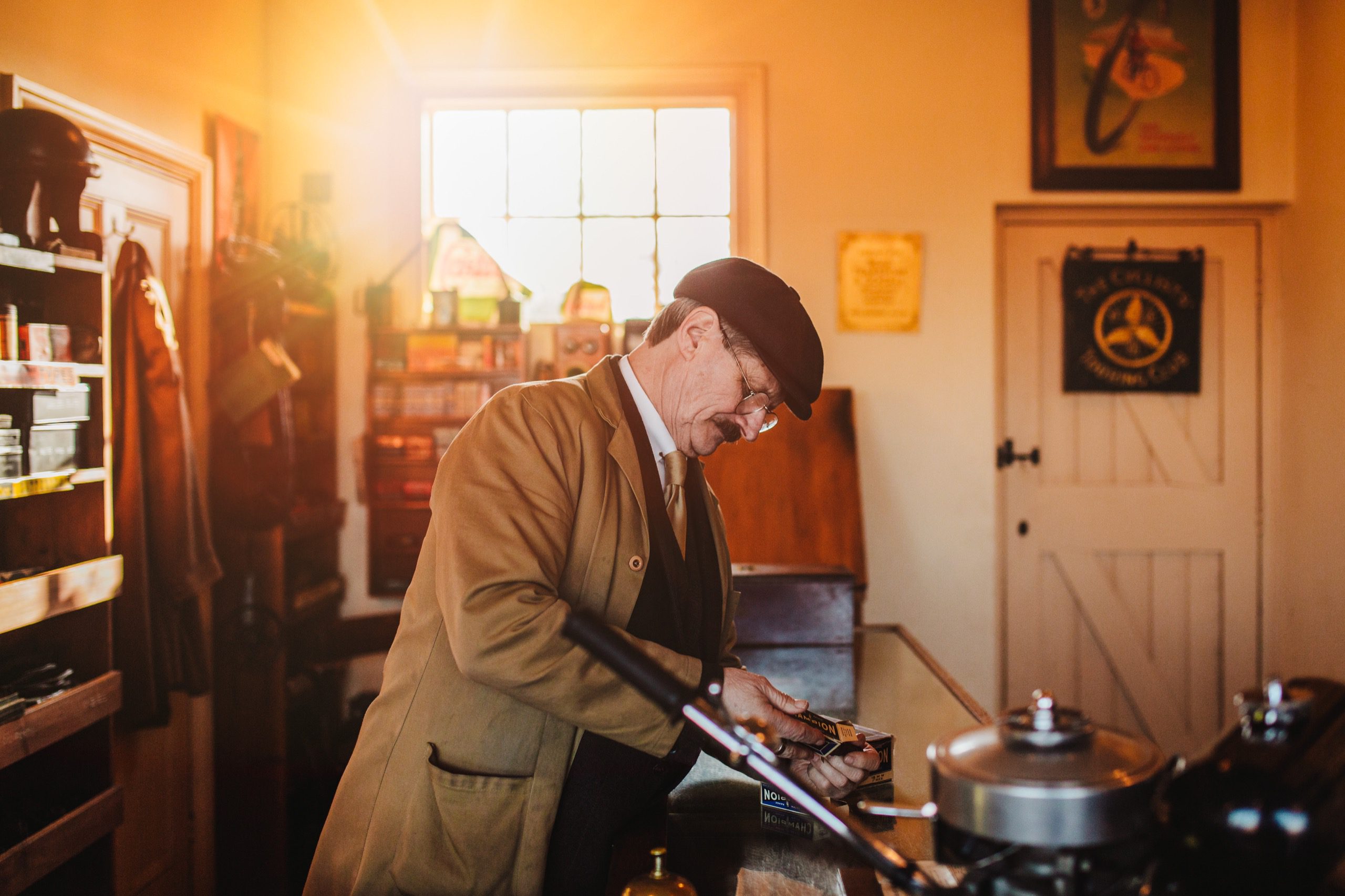 A man in a suit and tie, wearing a flatcap and brown jacket, in a workshop.