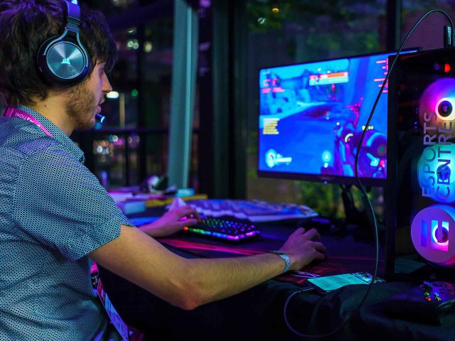 A PC gamer playing a game with a headset on, next to a computer tower with rainbow coloured fans.