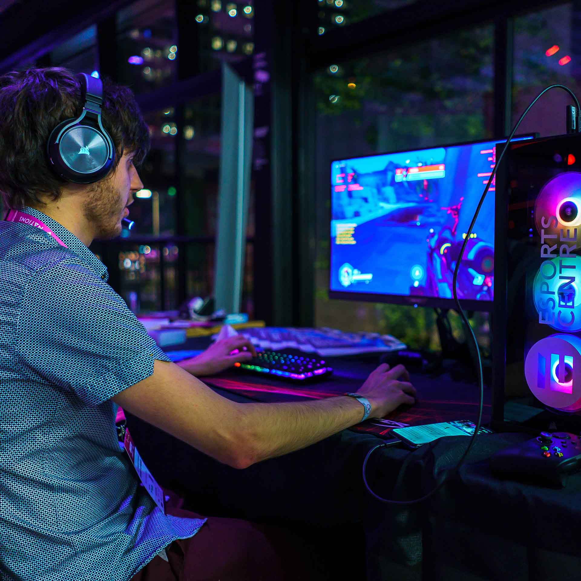 A PC gamer playing a game with a headset on, next to a computer tower with rainbow coloured fans.