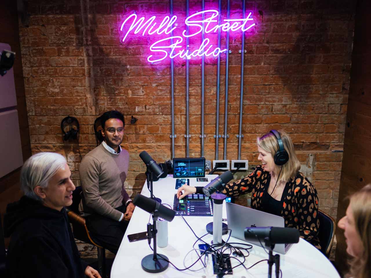 A recording studio with 4 microphones. Mill Street Studio is on the wall in pink neon lights.