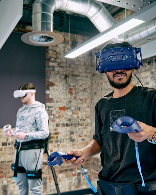 People wearing VR headsets and handheld controllers, strapped into sensors around their waists and legs.